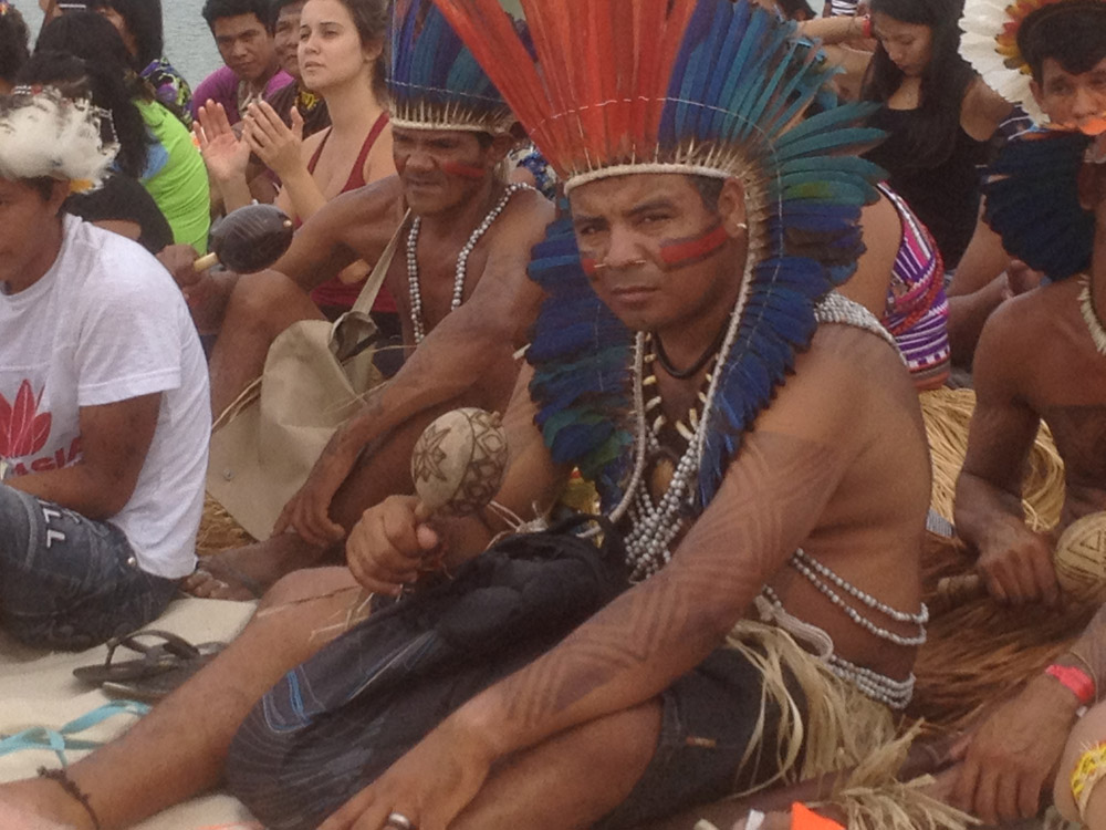 Indigenous man in red and blue feathered headdress sitting among a crowd shaking a rattle