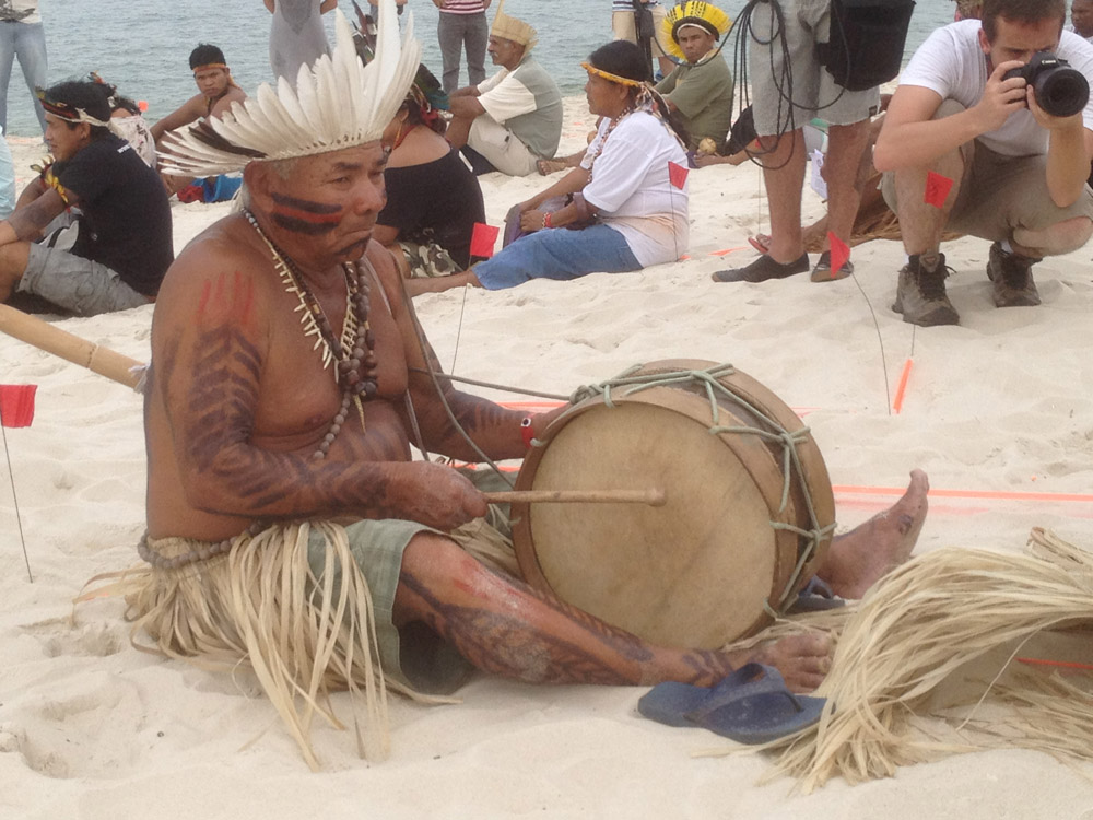Indigenous older man with white feather headdress sitting in the sand beating a large drum held between his ankles