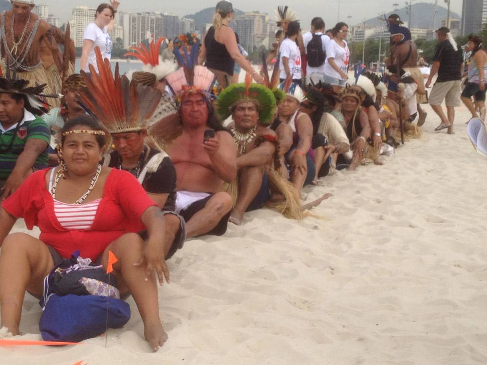 Indigenous people, some with feathered headdresses, sitting in a train line on the beach