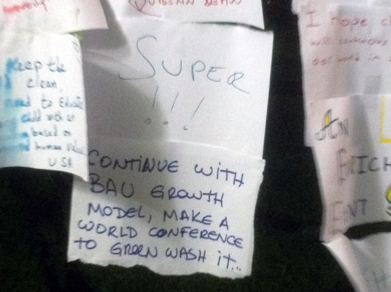 Handwritten slips of paper tacked to a clothesline. One in front reads, "Continue with BAU growth model, make a world conference to greenwash it."