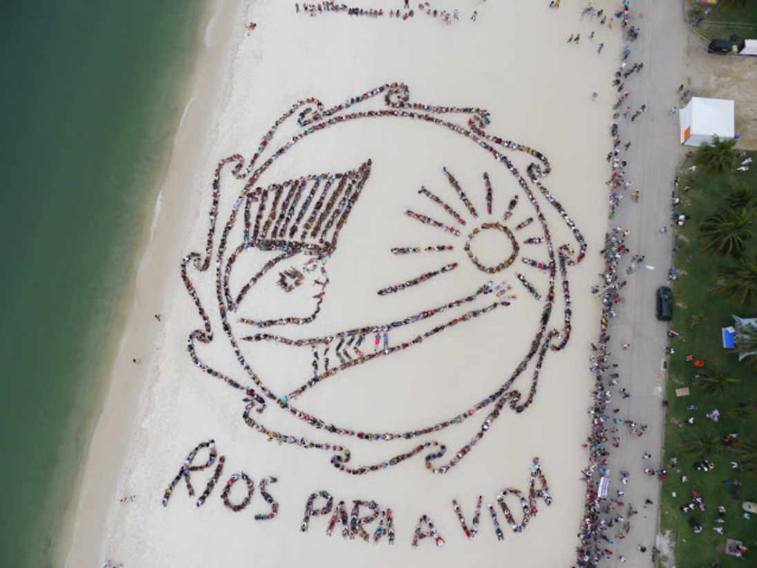 Drone shot of enormous picture in the sand: a huge circle drawn like waves around the head and arm of an indigenous person in a headdress reaching toward the sun, with the words "RIOS PARA A VIDA" spelled out below