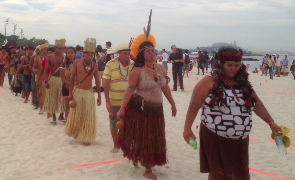 Indigenous people in traditional dress of grass skirts and headdresses filing onto the beach to take their places on the orange tape.
