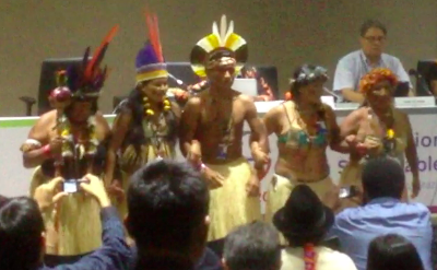 Indigenous people wearing traditional regalia of feather headdresses dance in front of a conference room