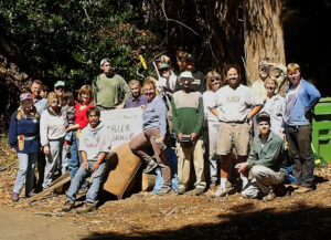 Group standing beside a bay tree in bright sunlight with a creek cleanup sign
