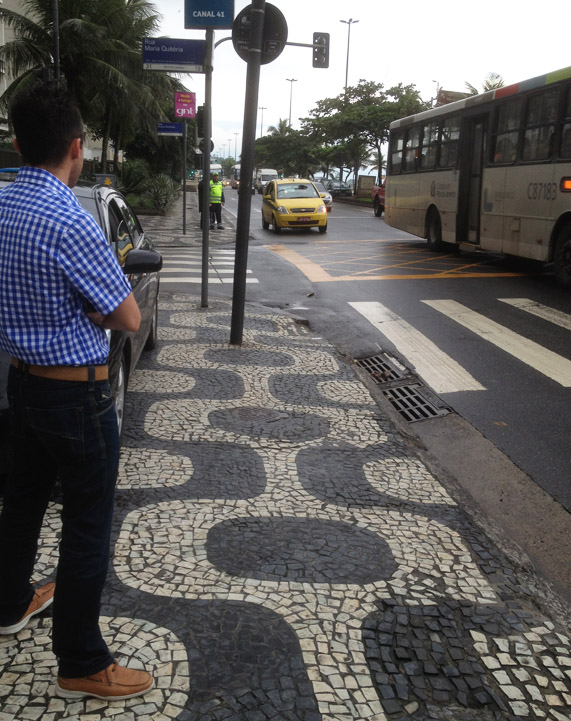 Rainy street with people waiting at taxi stand and black & white mosaic geometric designs in the sidewalk.